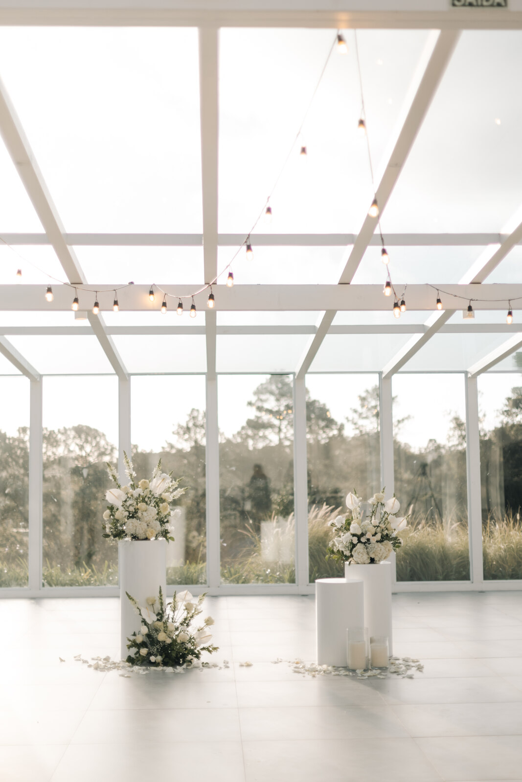 altar com trÊs colunas brancas com arranjos de flores brancas e velas dentro de salão branco e minimalista com janelas de vidro