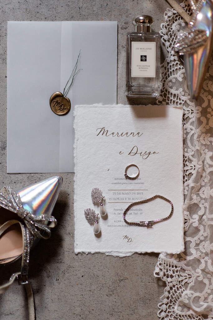 convite de casamento branco rústico com brincos aliança e pulseira em cima a lado de vidro de perfume e sandália prata