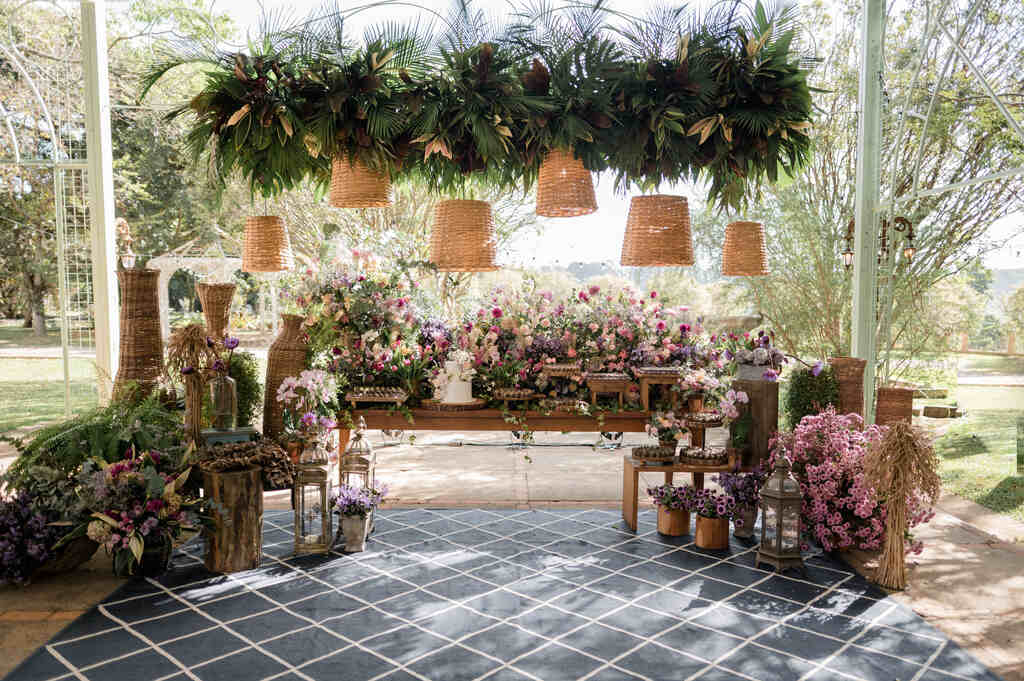 salão ao ar livre com mesas de madeira com bolo de casamento branco e ao lado arranjos com flores rosas roxas e lavandas