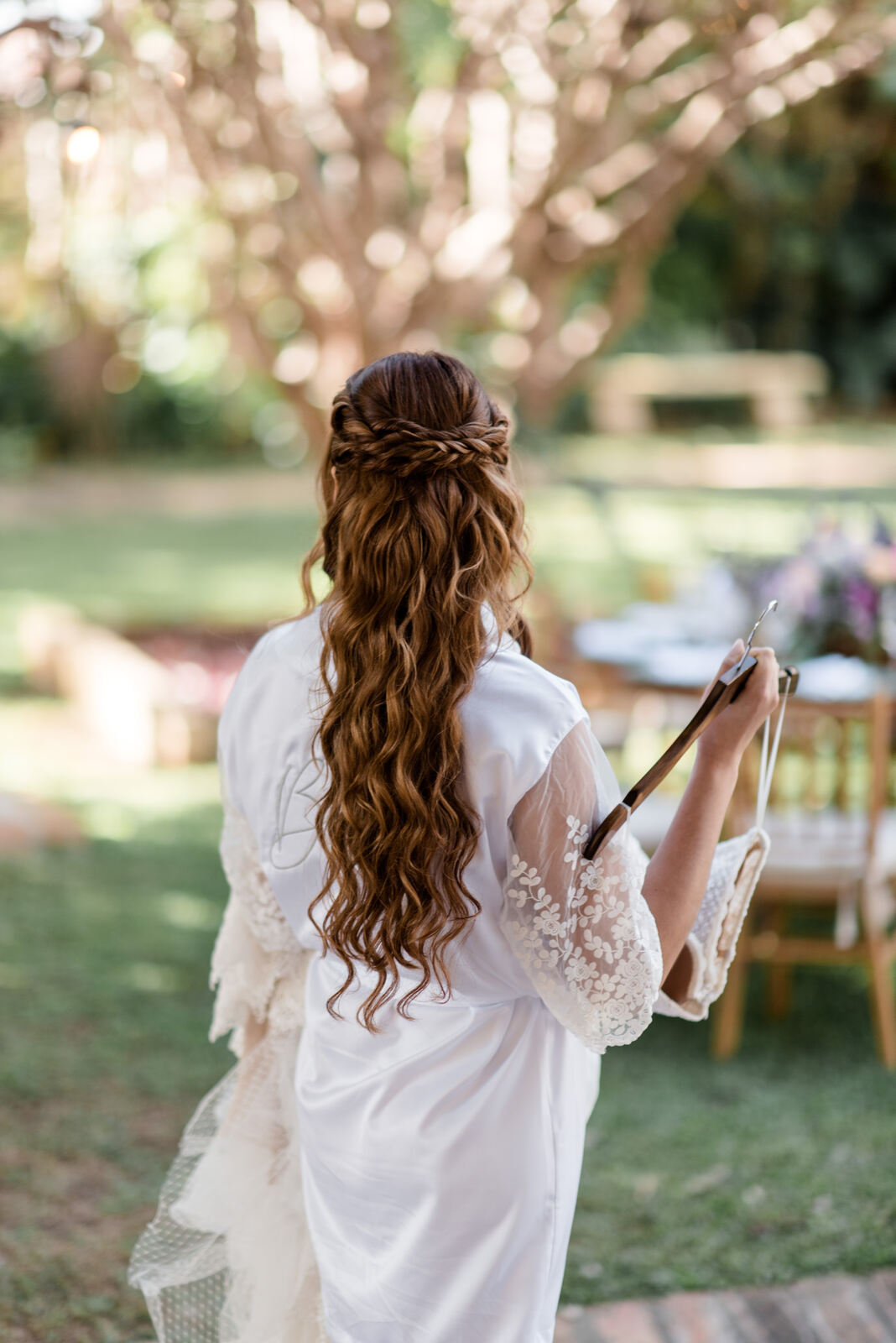 noiva com robe branco rendado de costas mostrando cabelo semi preso ondulado com coroa com tranças