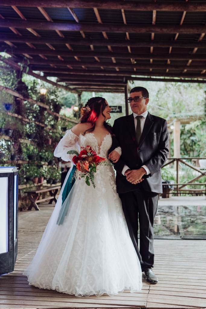entrada da noiva segurando buquê vermelho com laço verde ao lado do pai com terno preto