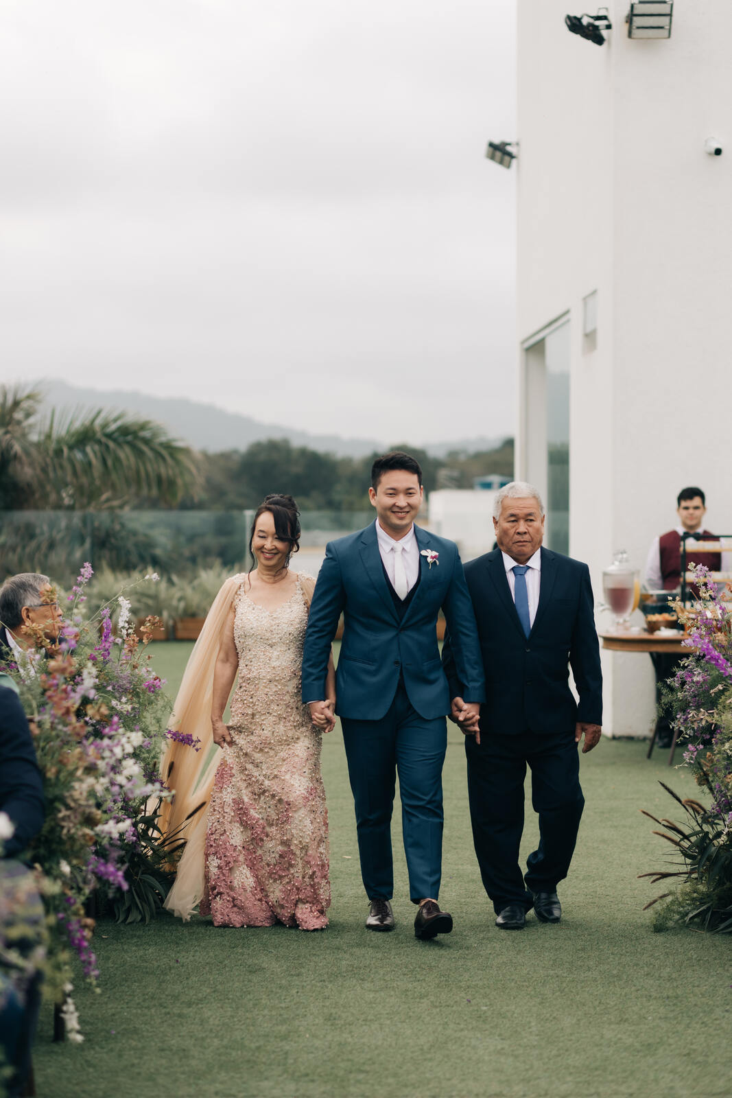 entrada do noivo com terno azul colete e gravata branca slim de mãos dadas com pai com terno preto e gravata azul e mãe com vetsido nude e rosa com capa
