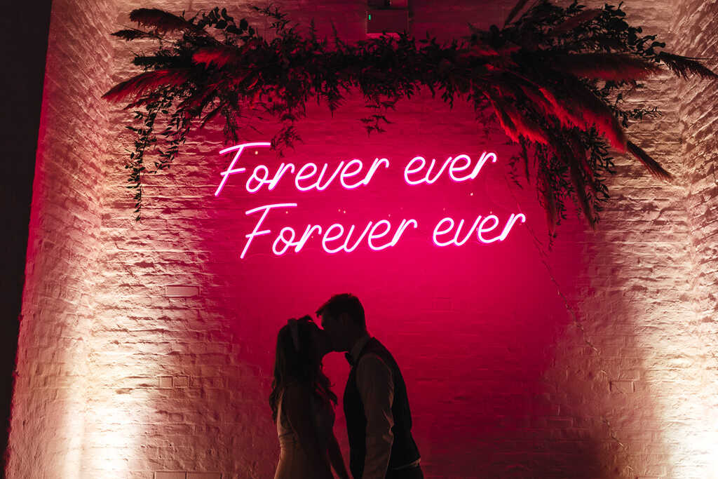 casal se beijando dentro de salão iluminado com letreiro neon escrito forever ever na cor fucsia