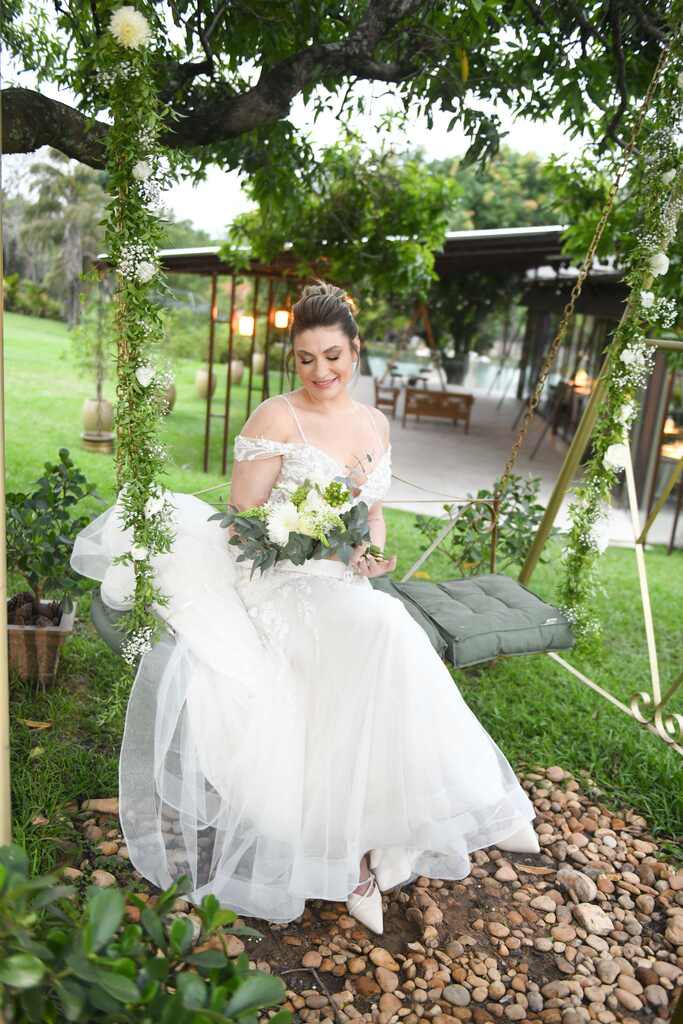noiva com topee segurando buquê com flores brancas sentada no balanço ao ar livre