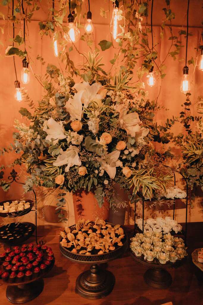 mesa com bandejas com doces de casamento e vaso com flores brancas e laranjas ao fundo