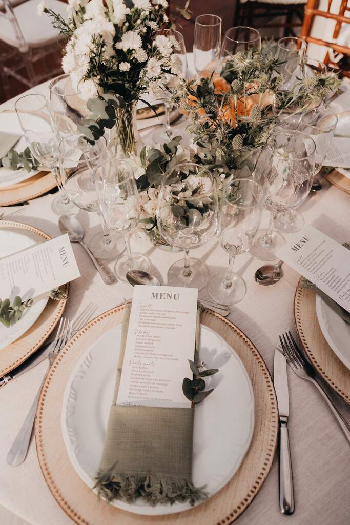 vista de cima de mesa posta com toalha branca com souplat bege e guardanapo verde com menu de casamento e ao centro taças e flores brancas e rosas laranjas
