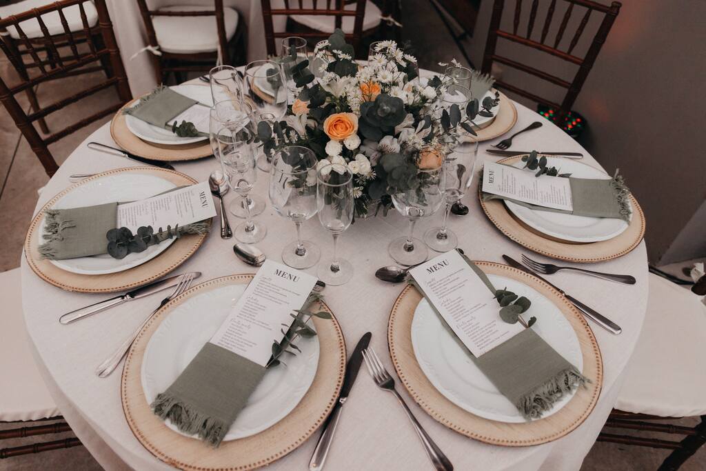 vista de cima de mesa posta redonda com toalha branca com souplat bege e guardanapo verde com menu de casamento e ao centro taças e flores brancas e rosas laranjas