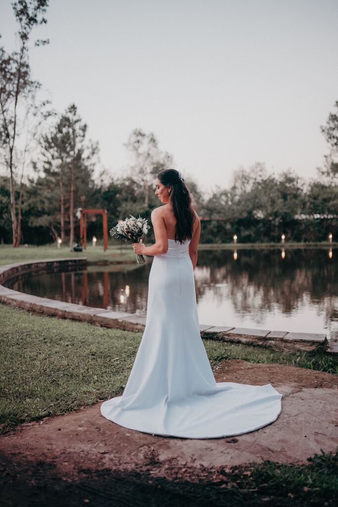 noiva de costas com cabelo semi preos e vestido minimalista sem alças segurando buquê com flores brancas no campo perto do rio