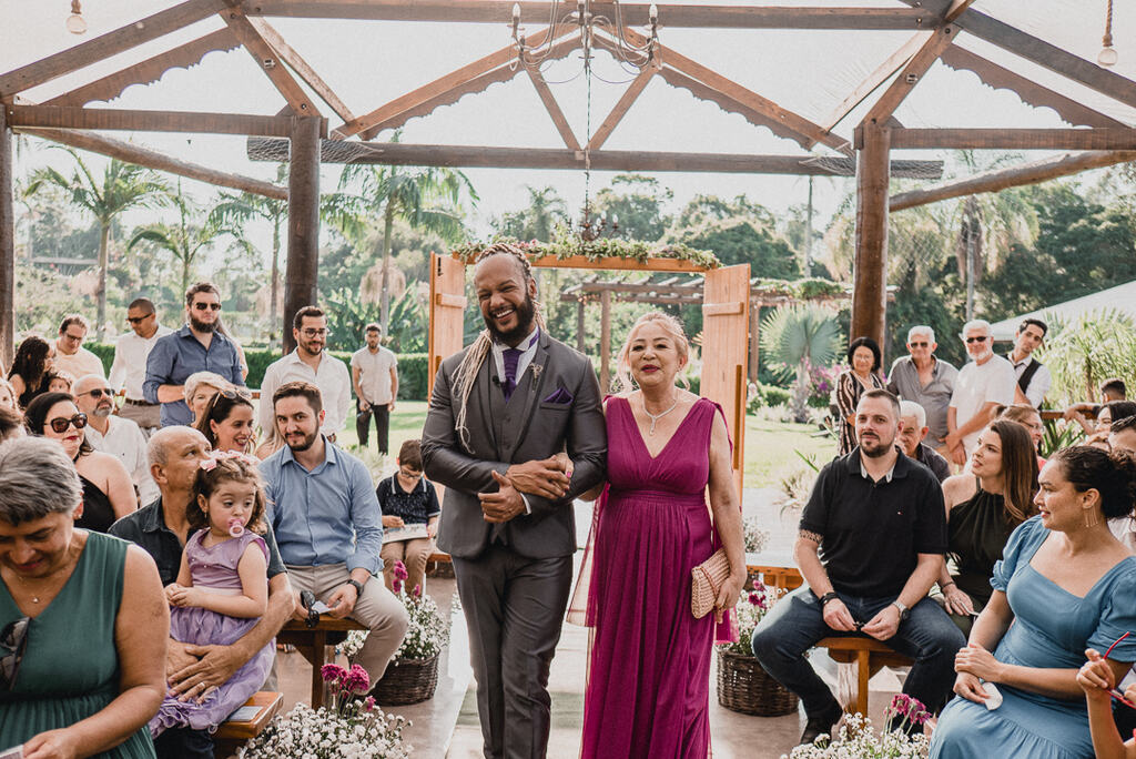 entrada do noivo com tenro cinza e gravata roxa ao lado da mãe com vestido na cor fúcsia