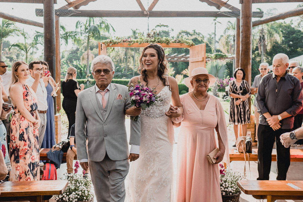 entrada da noiva com mini buquê roxo lilás e fúcsia ao lado da mãe com vestido e chapéu rosê e pai com terno cinz e gravata rosê