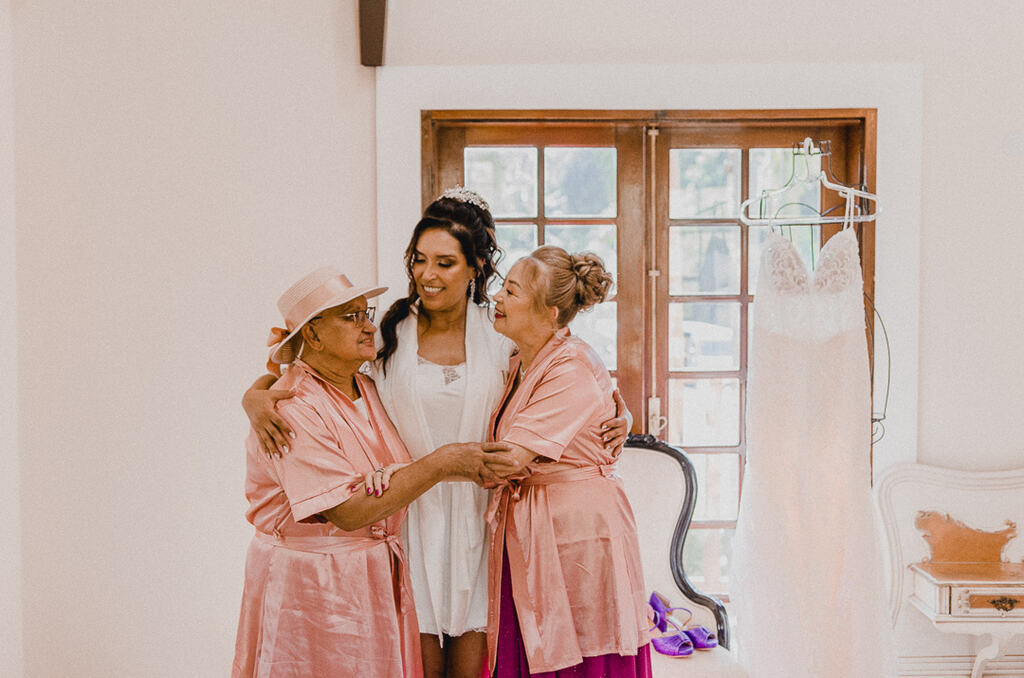 noiva com robe branco abraçando mãe e sogra com robê rosa claro dentro do quarto