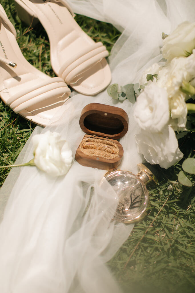 porta aliança de madeira sob véu de noiva vidro de perfume e sandália branca ao lado