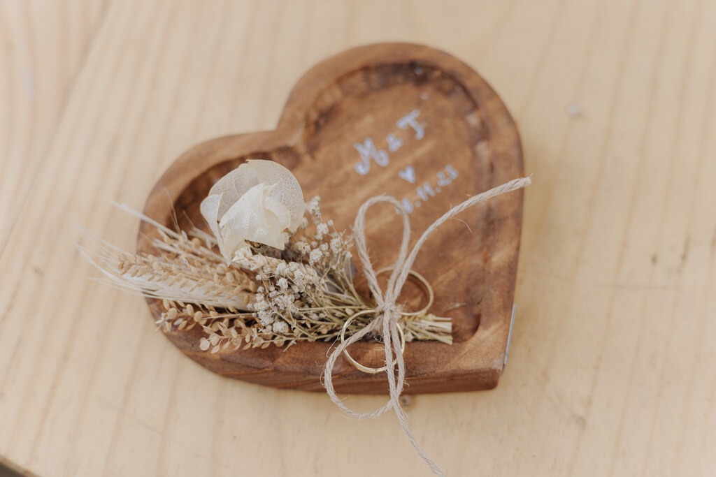 porta aliança de madeira em formato de coração com mini flor e laço prendendo alianças