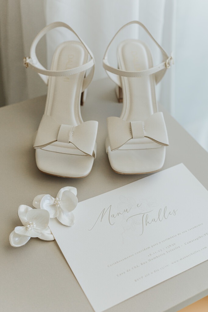 sandália branca para noiva com laço e convite de casamento ao lado de grinalda delicada em formato de orquidea