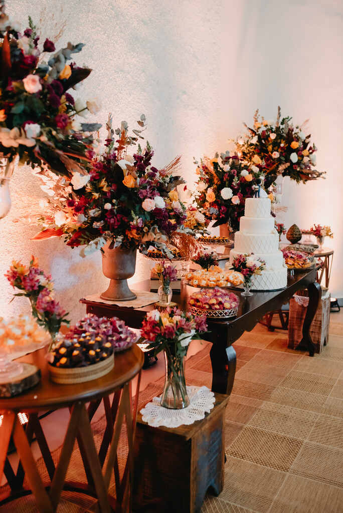 mesa com bolo de casamento branco e ao lado vasos com flores coloridas e bandejas rústicas com doces