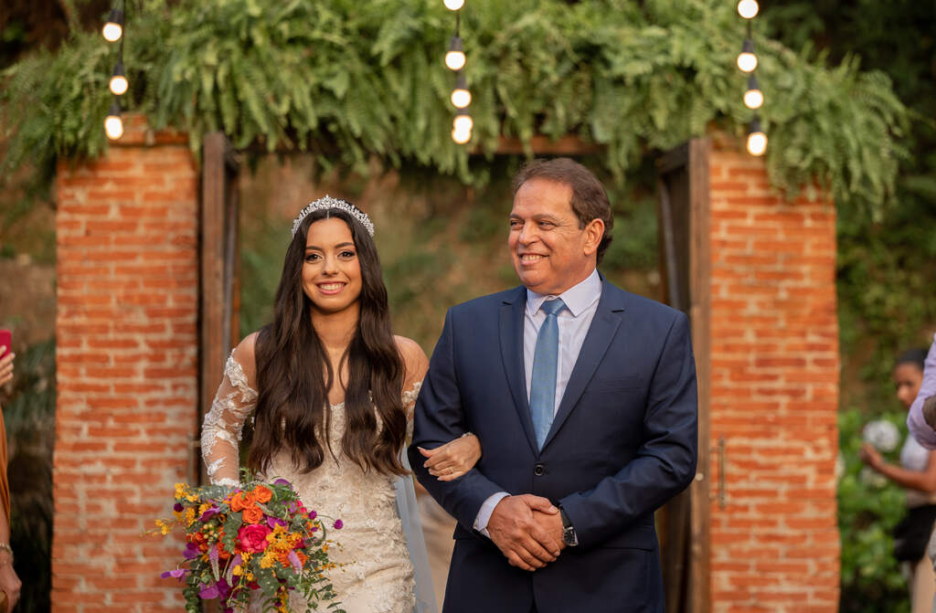 entrada da noiva com cabelo solto com tiara prateada segurando buquê colorido ao lado do pai com terno azul marinho e gravata azul média