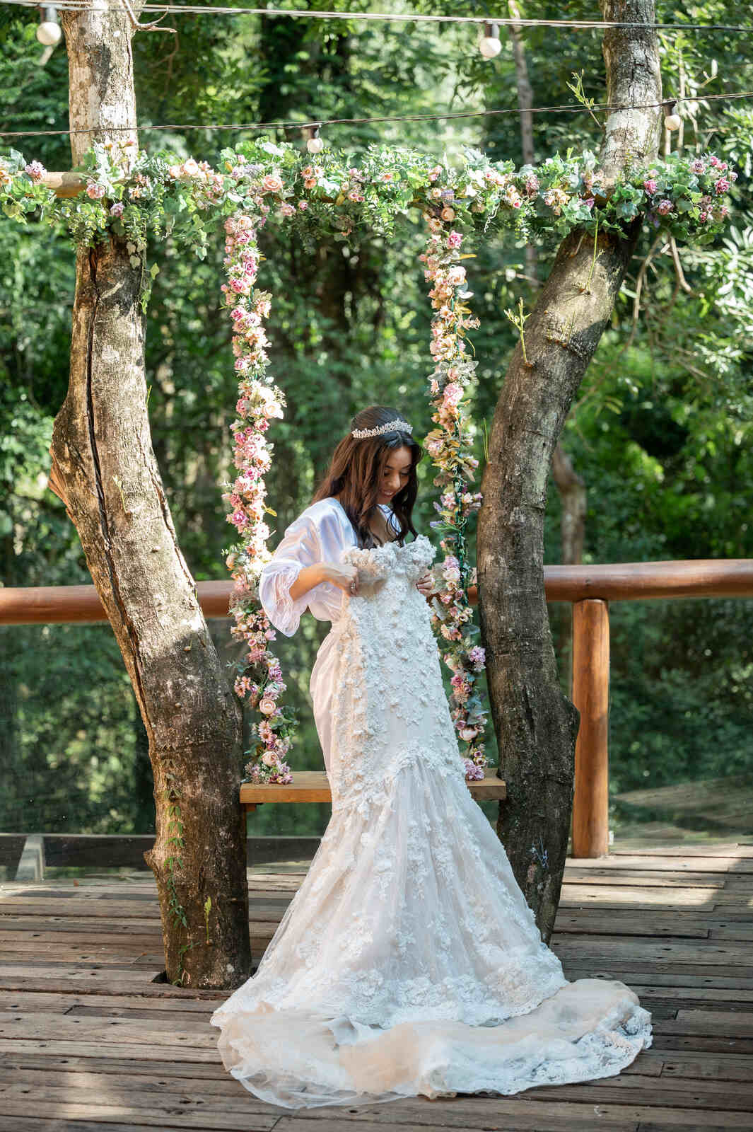 noiva com robe e tiara na varanda segurando o vestido de noivana frente de balanço rústico decorado com flores 