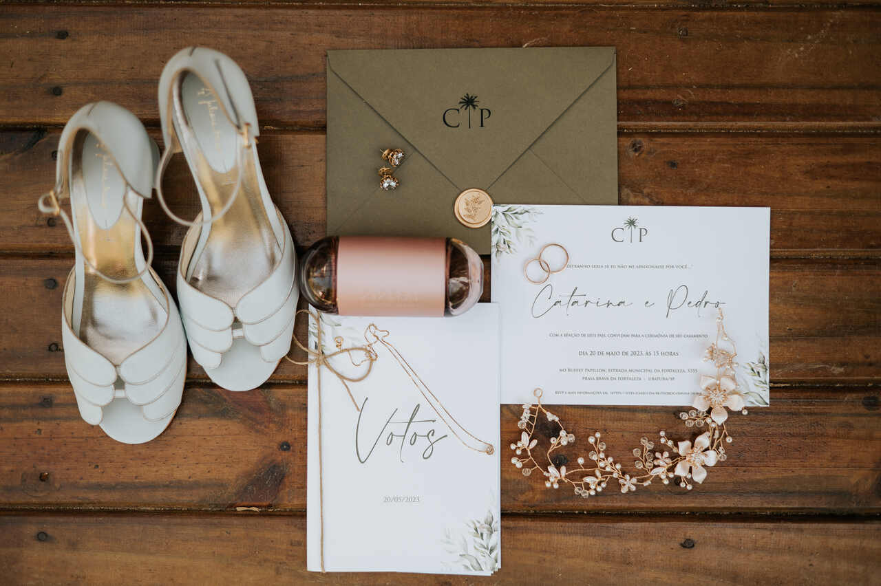 foto de cima do convite e caderno de votos branco com estampa de folhagens envelope marrom e ao lado sandália de noiva branca perfume 212 rosa e grinalda floral