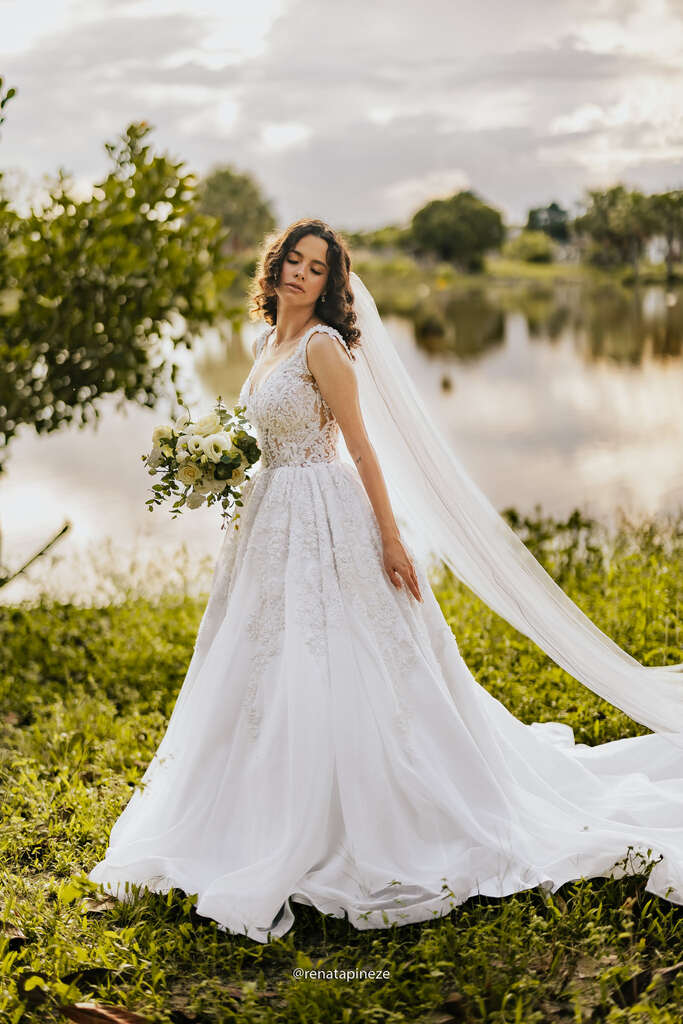 mulher com vestido de noiva com saia volumosa e véu longo segurando buquê com rosas brancas na frente de lago