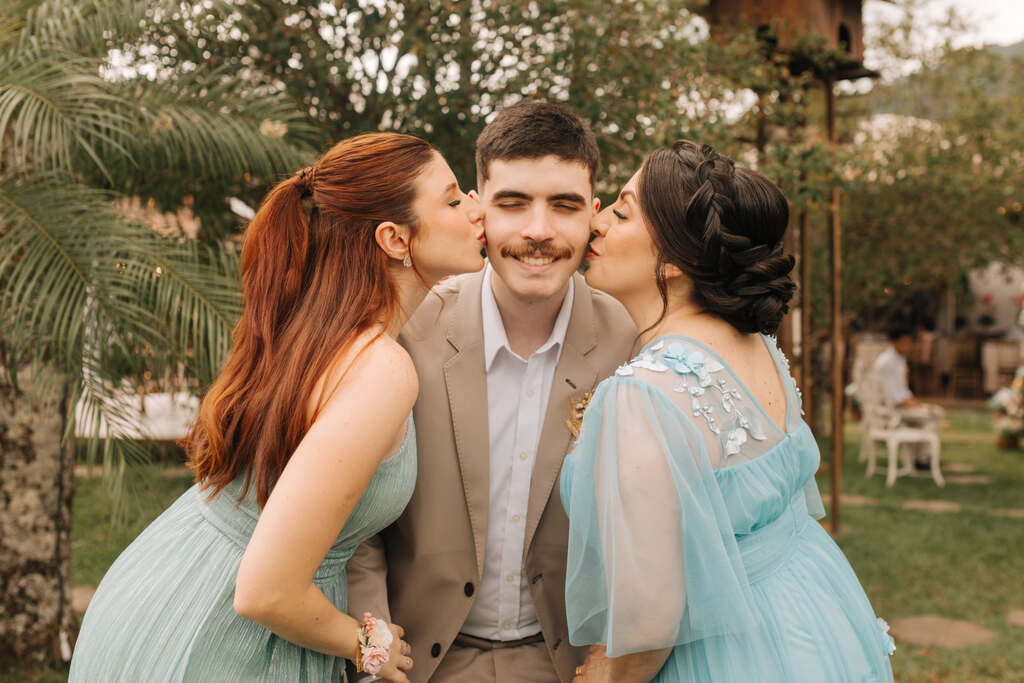 duas mulheres com vestido azul serenity beijando o rosto do noivo com terno bege claro