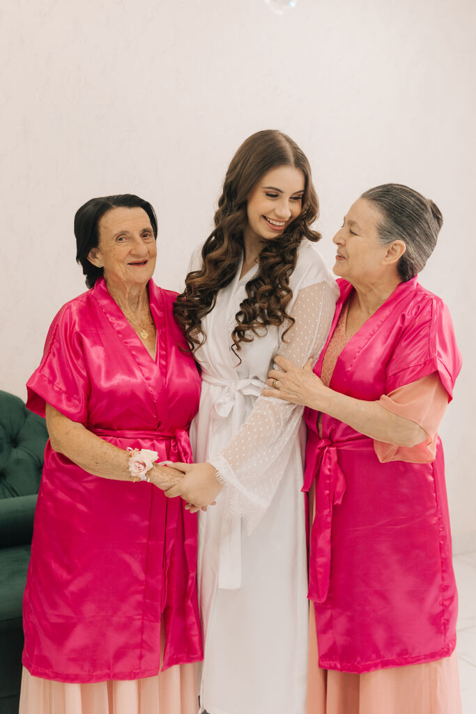 noiva com robe branco rendado ao lado de duas ssenhoras com robes na cor pink