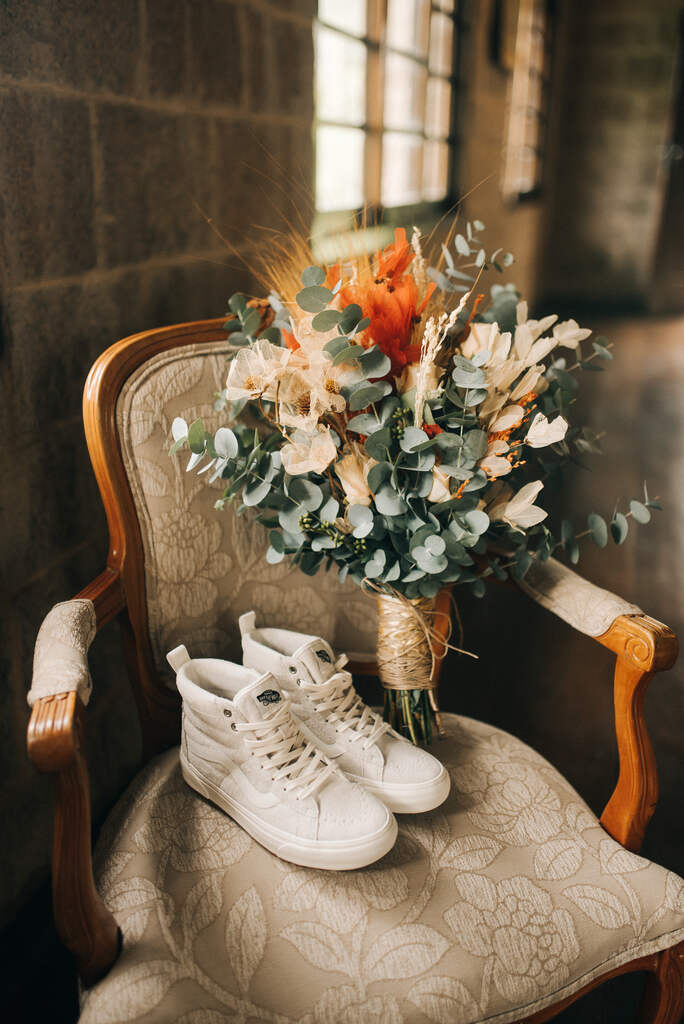 tênis de cano alto branco da noiva ao lado de buquê com ramos de eucalipto e flores laranjas e desidratadas