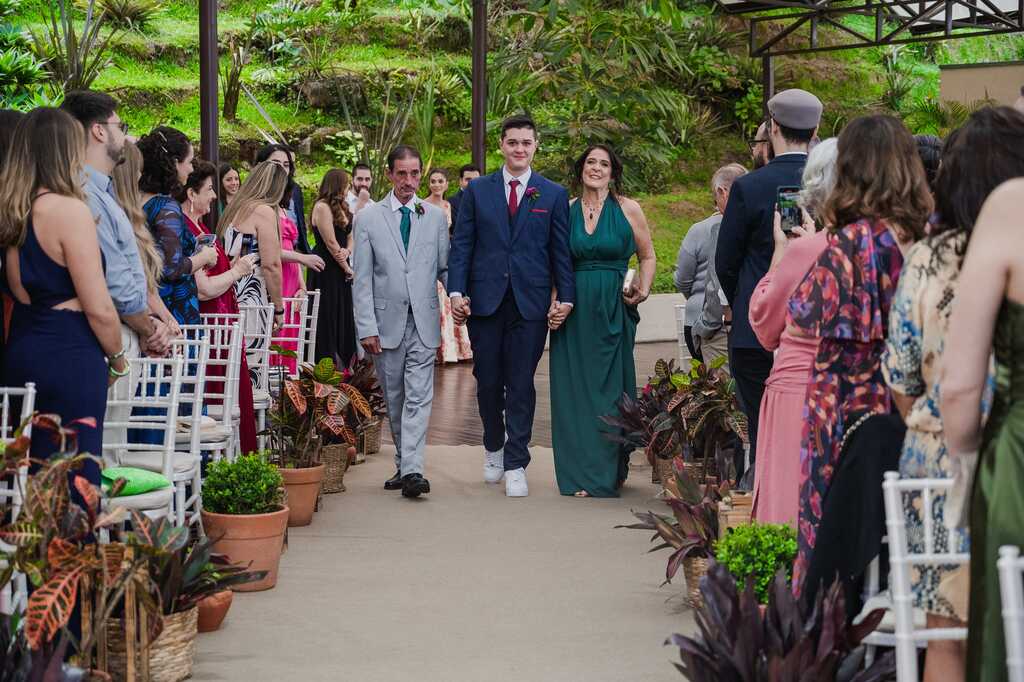 entrada do noivo com terno azul ao lado do pai com tenro cinza claro e mãe com vestido verde escuro