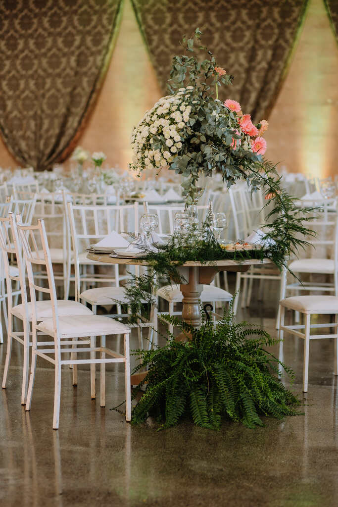 salão mesas comunitárias com cadeiras brancas e mesa decorada com plantas e vaso com flores amarelas e rosas