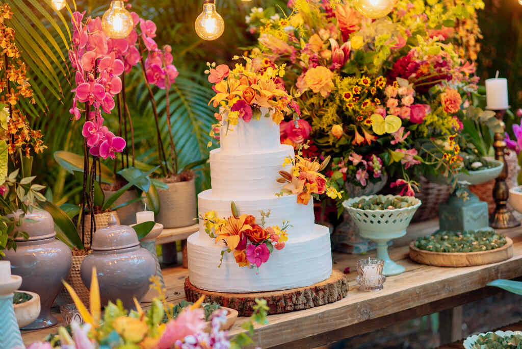mesa com bolo de casamento branco com três andares com flores amarelas e laranjas com flores e decoração tropical