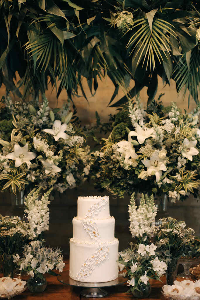 mesa de bolo de casamento branco com três andares e ao lado arranjos com flores e lírios brancos e arranjo aéreos com plantas