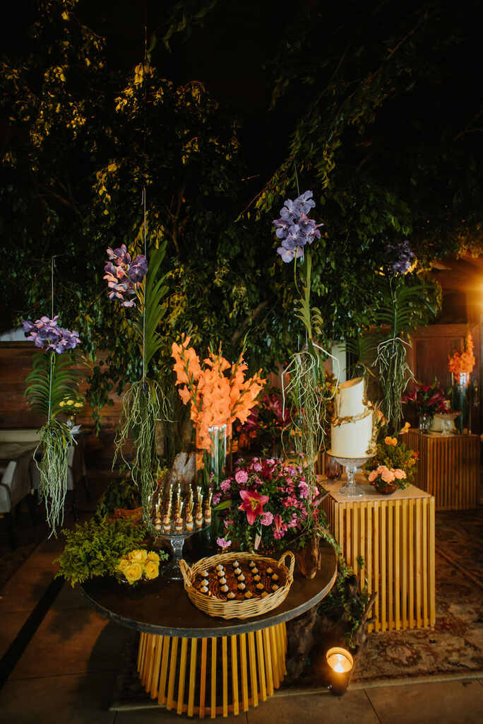 salão com mesa de doces de casamento em bandejas e ao fundo folhagens e flores azuis rosas e liláses