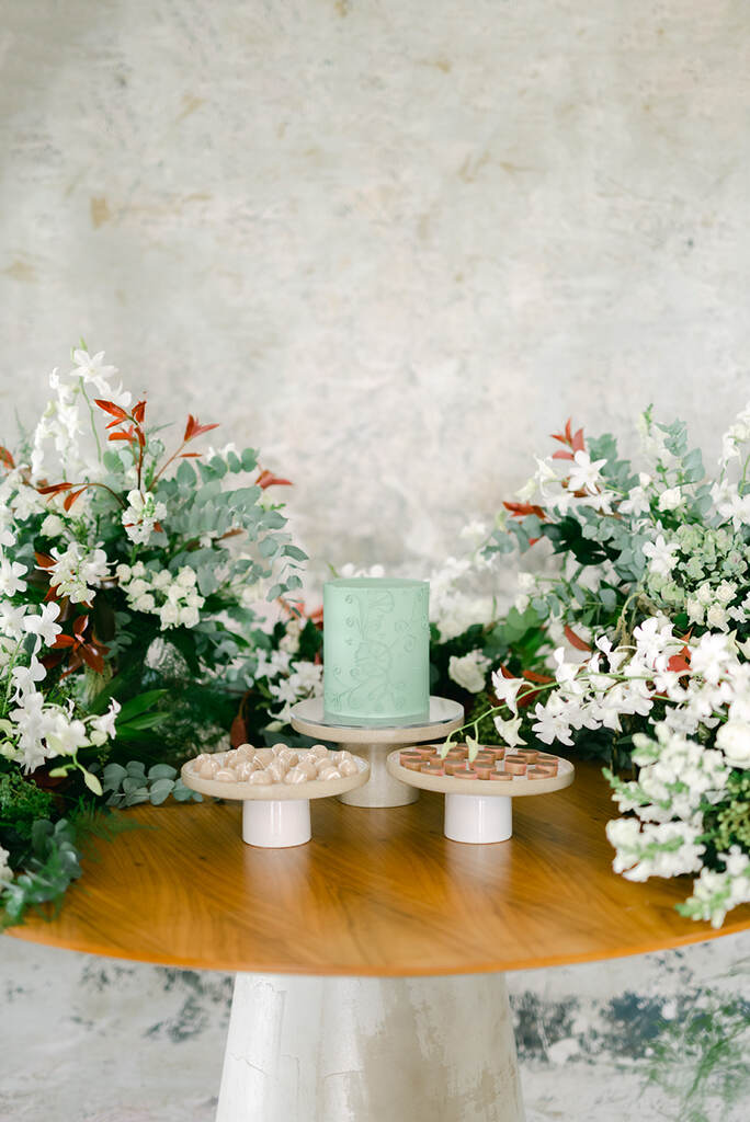 mesa com duas bandejas brancas com doces de casamento e bolo verde ao fundo e aos lados flores brancas e vermelhas