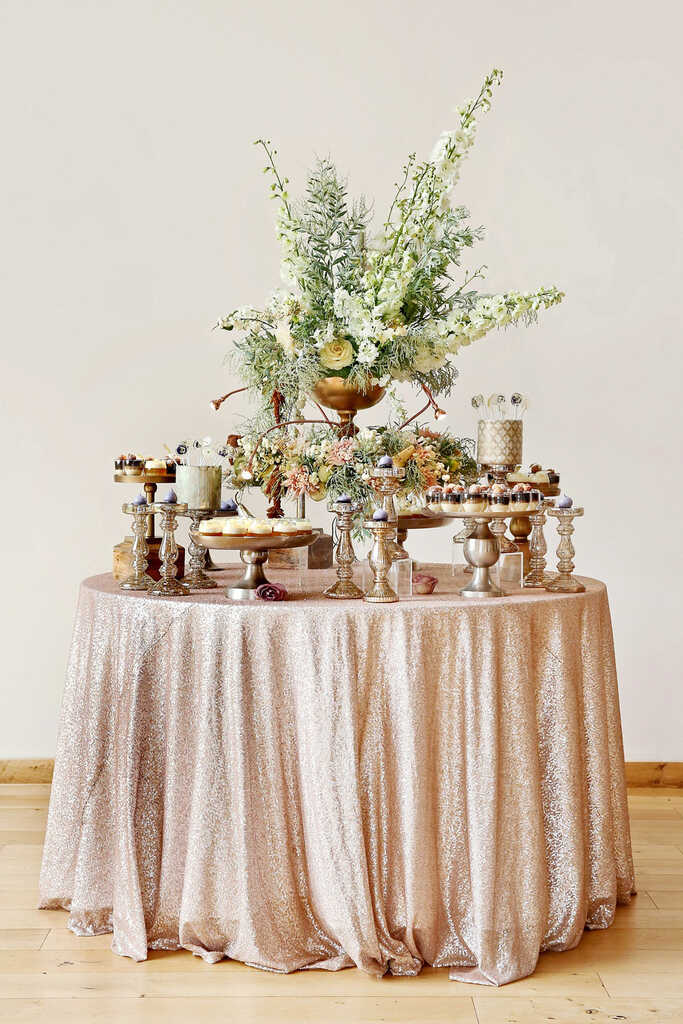 mesa de casamento com doces com toalha brilhante e arranjos com folhagens