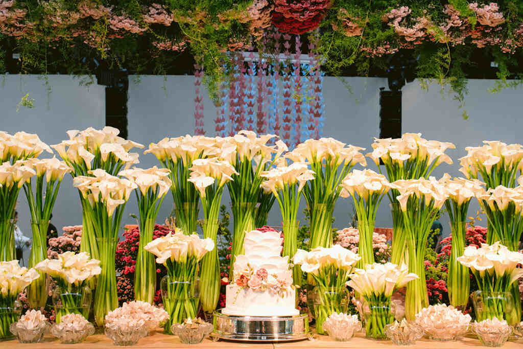 mesa com bolo de casamento com bandejas de vidro com doces de casamento e ao fundo vasos com tulipas brancas