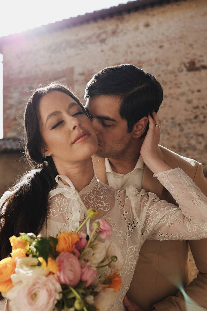 noivo beijando o rosto da noiva enquanto ela segura buquê com flores brancas laranjas e rosas