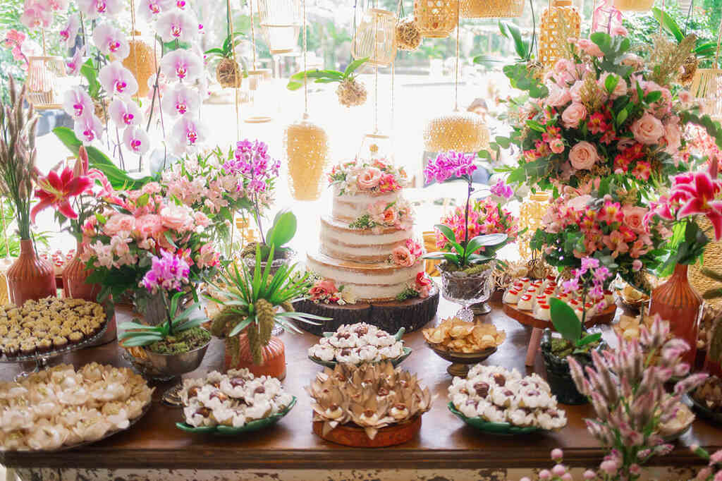 Mesa com bolo de casamento semi espatulado com topo de flores e ao redor bandejas com docs e flores cor de rosa e lilas