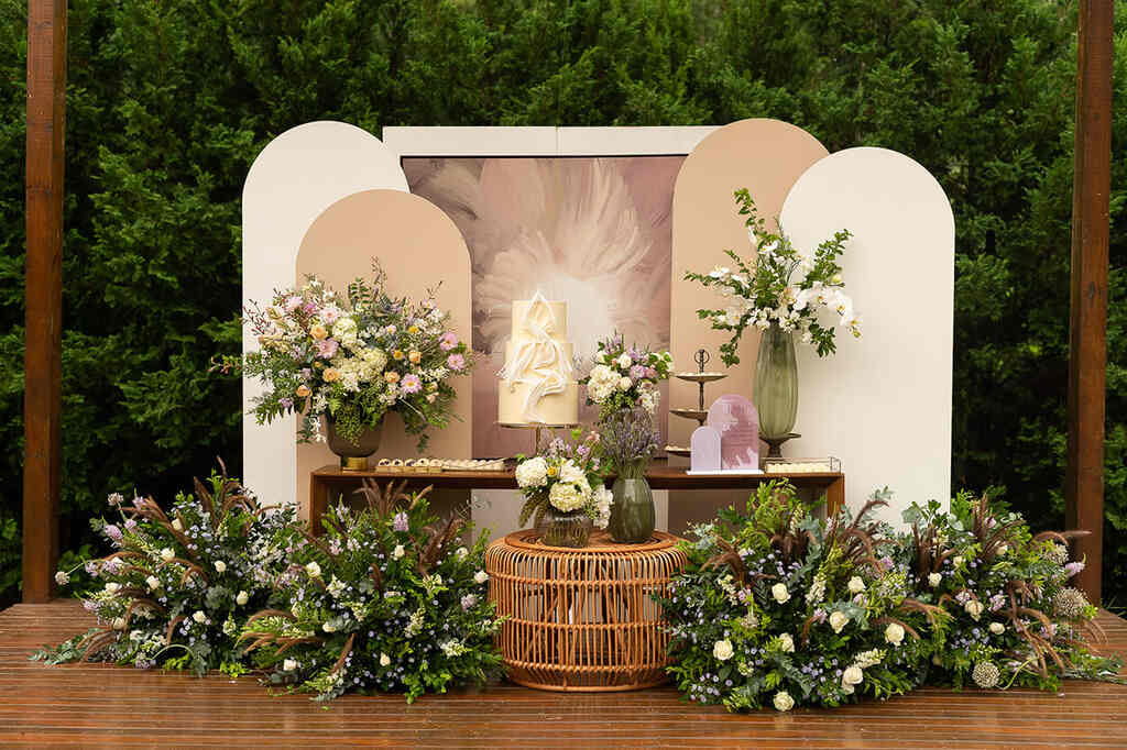 Mesa de bolo de casamento com decoração moderna com paineis arredondados na cor bege e off white e arranjos com flores lilás amarela e rosa