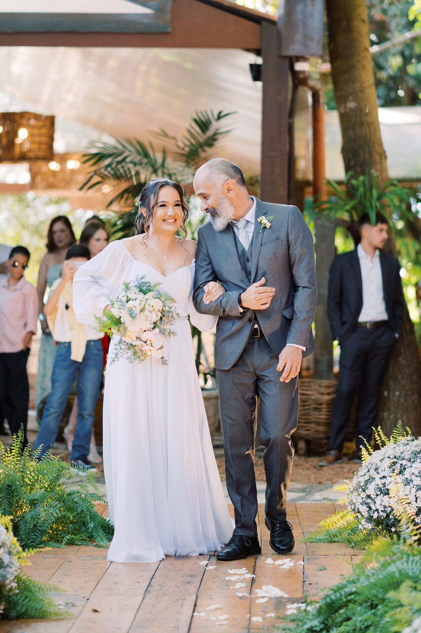 mulher com vestido de noiva minimalista com manga longa segurando buquê branco entrando com o pai com terno cinza