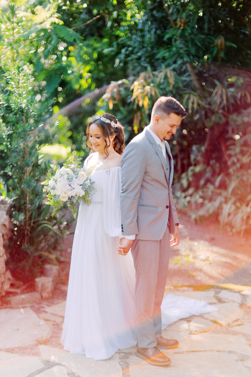 mulher com vestido de noiva minimalista com manga longa segurando buquê branco de mãos dadas durante o first touch com o noivo com terno cinza