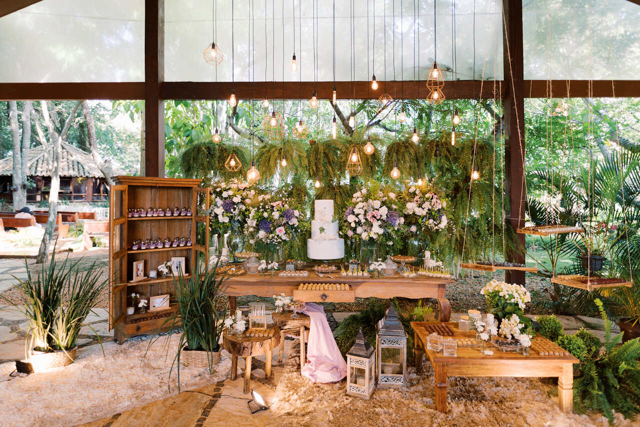 salão coberto com mesa rústica para bolo de casamento ao lado com vasos com flores brancas roxas e lavandas