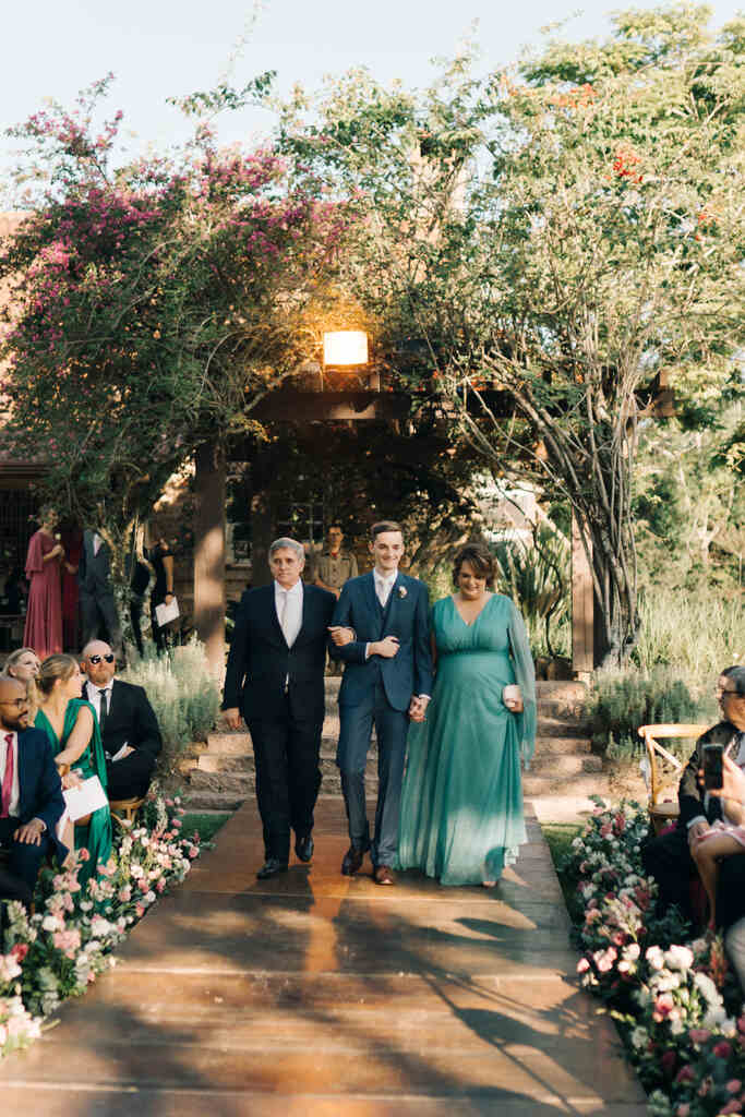 entrada do noivo com terno e colete azul e gravata branca ao lado do pai com terno preto e mãe com vetsido verde esmeralda
