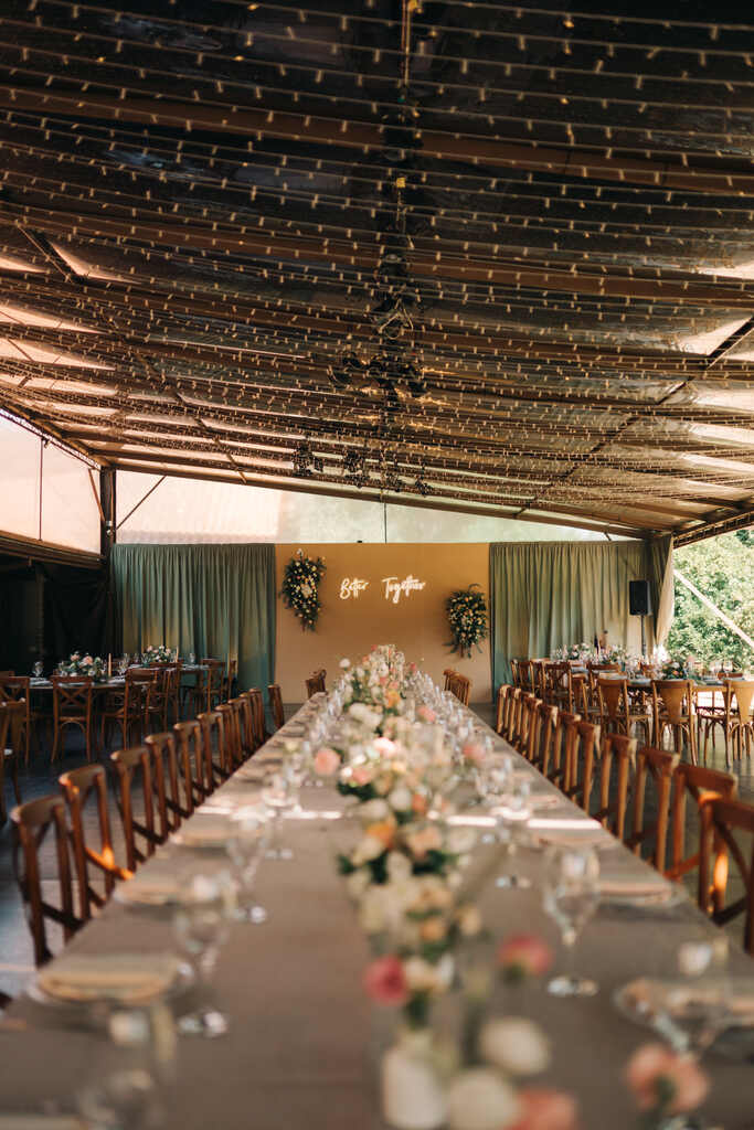 salão com mesa comunitária posta com flores no centro e letreiro neon ao fundo