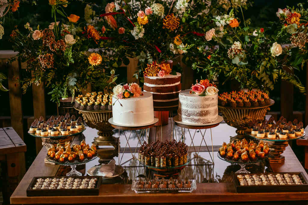 mesa com três bolos de casamento decorada com flores e ao lado bandejas de vidro e madeira com doces de casamento