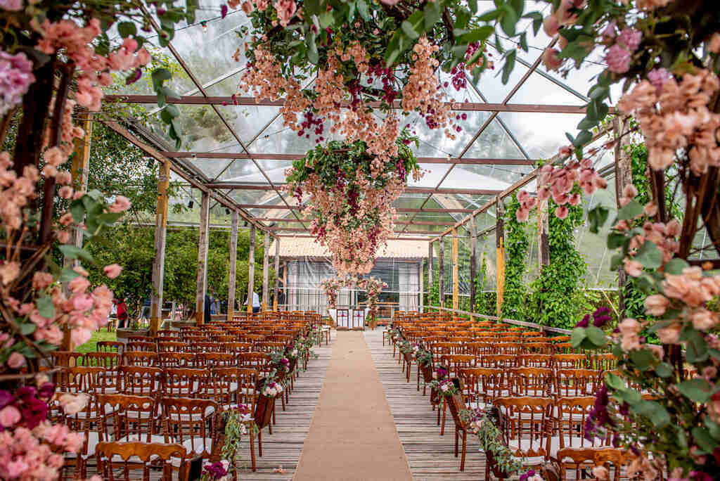 espaço com decoração para casamento de dia com capela de vidro com cadeiras de madeira e flores cor de rosa