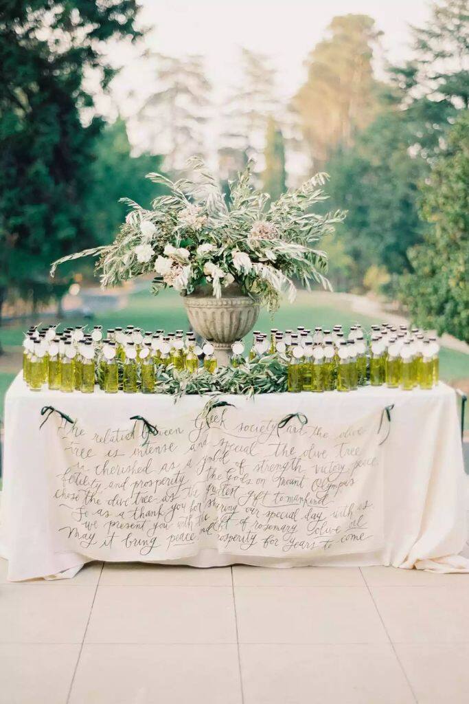 decoração de casamento de dia com mesa com toalha branca ao ar livre com vaso com flores brancas no centro e vidros de azeite para lembrancinhas