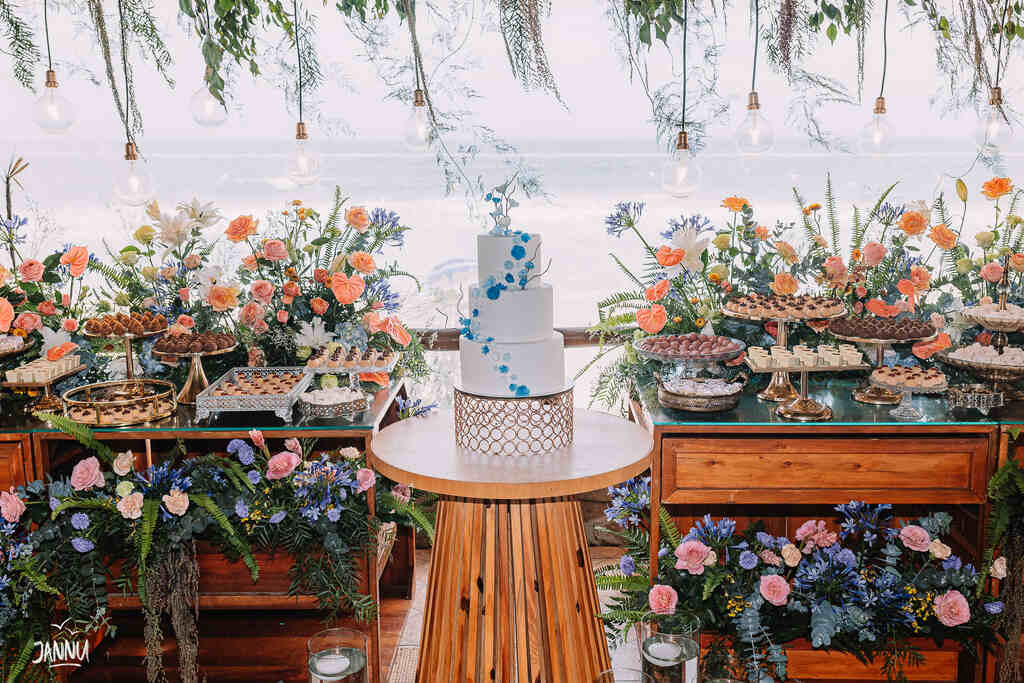 mesas de madeira com bolo de casamento branco com três andares com flores azuis e ao fundo mesas decoradas com flores e bandejas com doces