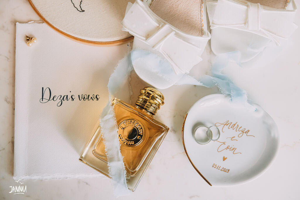 porta aliança branco de porcelana em formato de coração ao lado de perfume e sapato de noiva branco com laço