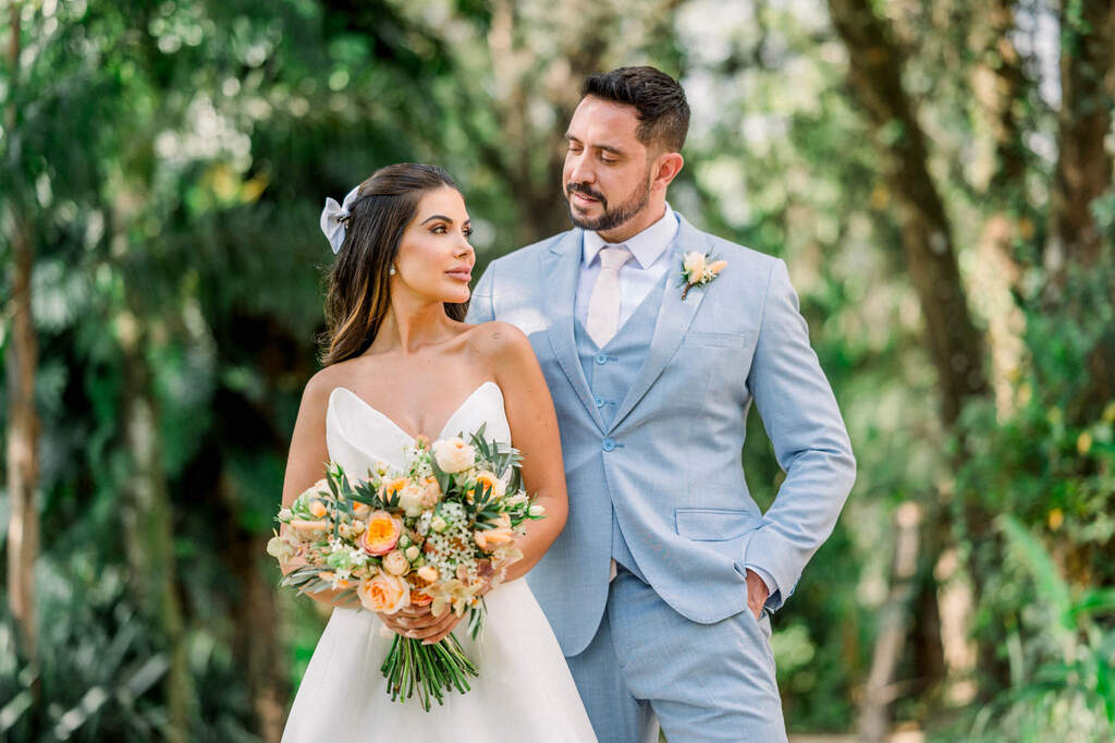 noiva com vestido sem alças segurando buquê com rosas laranjas ao lado do noivo com terno azul claro