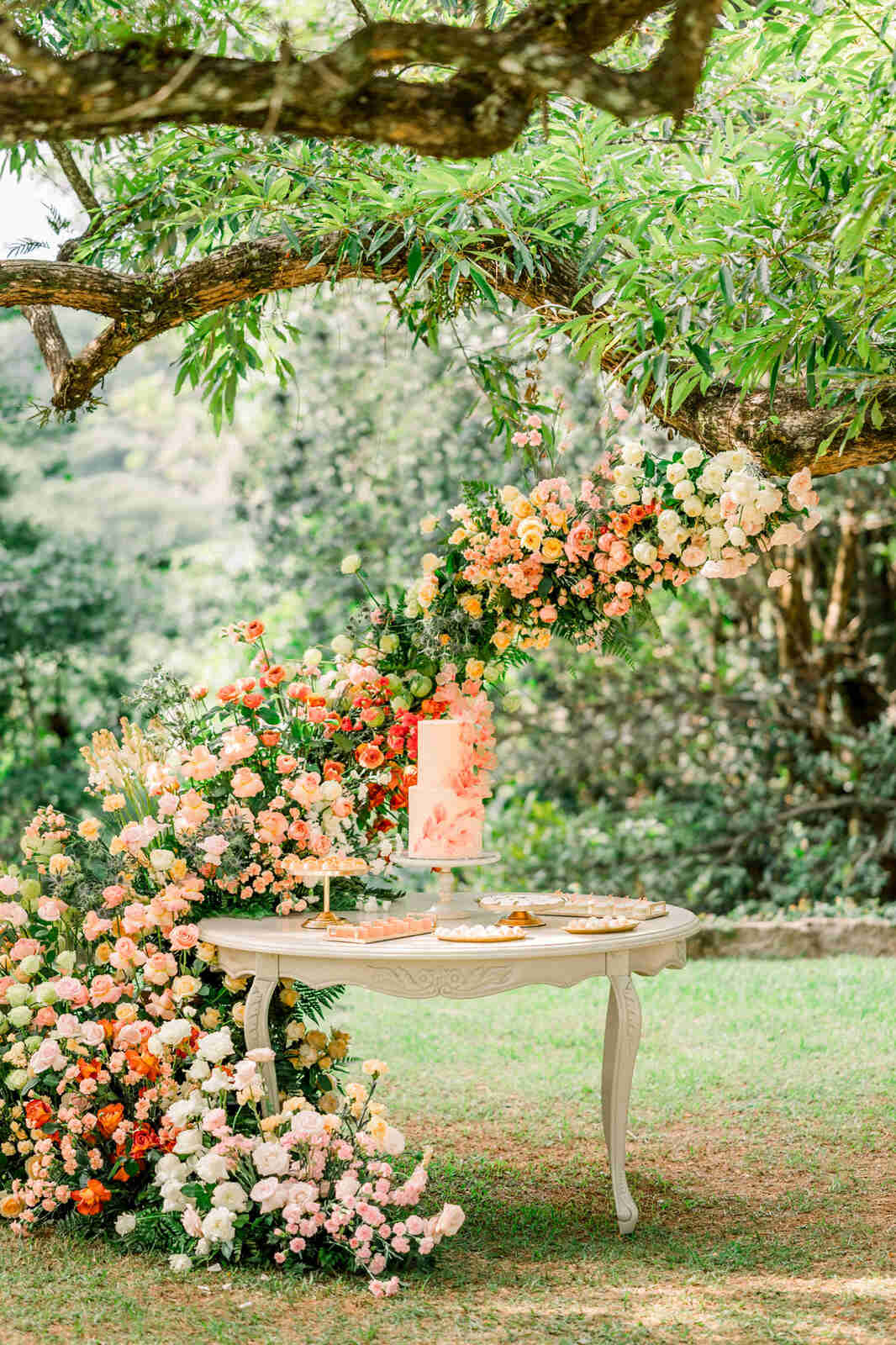 mesa redonda clássica com bolo de casamento na cor peach fuzz e ao fundo arco com flores brancas rosas e laranjas