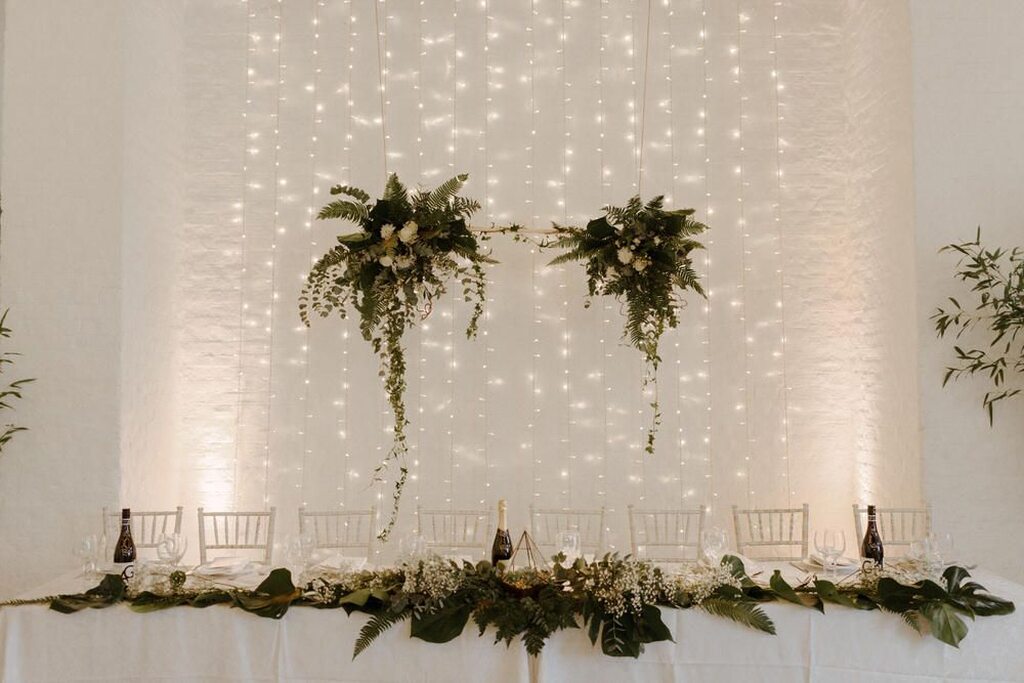 salão decorado com varal de luzinhas e mesa posta decorada com folhagens e flores brancas